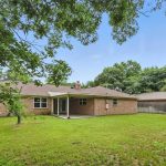 A single-story brick house with a covered patio in a spacious, grassy backyard, surrounded by trees and a wooden fence under a clear blue sky.