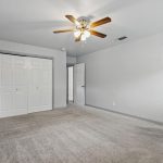 Spacious, empty bedroom with beige carpet, white walls, a ceiling fan with lights, double closet doors, two windows, and two open doors leading to other rooms.