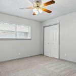 A simple, empty bedroom with light gray walls, beige carpet, a ceiling fan with lights, a window with blinds, and a closed double-door closet.