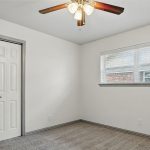 A small empty room with white walls, gray carpet, a double-door closet, a window with blinds, and a ceiling fan with lights.