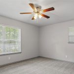 A simple, empty room with light gray walls, carpeted floor, two windows with white blinds, and a ceiling fan with wooden blades and a light fixture. Natural light enters through the larger window.