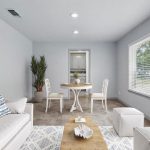 A bright living room with light gray walls, a white sofa with blue and white pillows, a wooden coffee table, two white ottomans, a round table with two chairs, a large plant, and a big window letting in natural light.