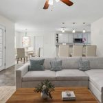 A modern living room with a large L-shaped sofa, wooden coffee table, and wall art. In the background, there’s a dining area and a kitchen with pendant lights. French doors lead to a green backyard.