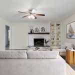 A modern living room with a white couch, armchair, and wooden side table facing a white brick fireplace. Built-in shelves with decor, large windows, sailboat paintings, and a ceiling fan complete the space.