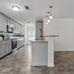 Modern kitchen with gray cabinets, stainless steel appliances, a tiled backsplash, and marble countertops. Pendant lights hang over a breakfast bar, and large floor tiles cover the spacious open area.