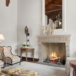Elegant living room with a lit fireplace, ornate mantel, two patterned armchairs, artwork, wooden side tables with lamps, and high ceilings with a stained glass window. Soft, neutral color palette throughout.