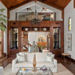 Spacious living room with high, wood-beamed ceiling, stained glass windows, large indoor plant, white sofa, glass coffee table, tapestry on the wall, and decorative sculptures with a balcony above the entryway.