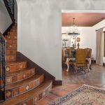 A tiled staircase with ornate wrought iron railing leads to an open dining room with a chandelier, wooden table and chairs. The entryway has a patterned rug, decorative pillar, and framed wall art.
