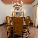 Elegant dining room with a dark wood table, six brown leather chairs, a crystal chandelier, large windows with beige curtains, and a mirror above a sideboard. The floor is hardwood and the ceiling is painted a warm color.