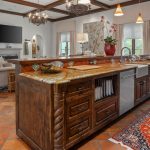 Spacious kitchen with a large wooden island, granite countertop, built-in sink, and dishwasher. Open living area with sofas, TV, chandeliers, tiled floor, and an orange patterned rug. Large windows brighten the room.