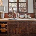 A small kitchen area with dark wood cabinets, a stainless steel sink, woven baskets, ceramic containers, and stacked plates on the countertop, beneath a window with sunlight streaming in.