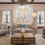 A cozy living room with a beige sofa, floral armchairs, a wooden coffee table, decorative pillows, books, large wall art of gold branches, and elegant chandeliers. The room has large windows and neutral decor.