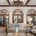 Elegant living room with exposed wood beams, five large arched windows, neutral-toned sofas, a floral armchair, tile flooring, and a decorative chandelier. Outdoor patio visible through the windows.