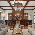 Elegant living room with exposed wood beams, two beige sofas with blue pillows, a chandelier, and a rustic wood kitchen with arched windows and bar stools. A cozy, inviting, and stylish space with warm lighting.