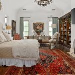 A cozy bedroom with a large bed, white bedding, vintage chairs, a red patterned rug, a fireplace, a wall-mounted TV, bookshelves, and large windows letting in natural light.