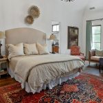 A cozy bedroom with a large bed, neutral bedding, two bedside tables with lamps, an ornate red rug, a vintage sofa with pillows, armchair, and paintings on white walls with natural light from windows.