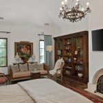 A cozy bedroom with a large bed, ornate chandelier, fireplace, wall-mounted TV, bookshelves, and a sitting area with a sofa, armchairs, and artwork. Natural light enters through two windows with green curtains.