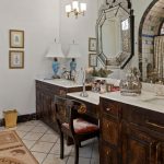 Elegant bathroom with dark wood cabinets, marble countertops, a decorative mirror, vintage lamps, framed art, and a large arched window overlooking a garden. A cushioned chair sits at a vanity area.