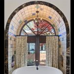 A large arched window with decorative tiles surrounds a white bathtub. Curtains are partly drawn, revealing a view of a red-leafed tree and a courtyard outside. A chandelier hangs above the bathtub.