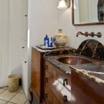Elegant bathroom with a dark marble countertop, vessel sink, large ceramic vases, a wooden vanity, wall-mounted faucet, mirror, and white walls. A soap dispenser and small towel are near the sink.