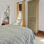 A cozy bedroom with a quilted bed, floral pillows, an antique green armoire, yellow curtains, large windows, a mirror, and a view into a bathroom with plants on the sink.