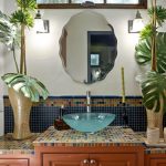 A bathroom vanity with a colorful mosaic countertop, a glass vessel sink, two large vases with tropical leaves, a round mirror, wall lights, and green hand towel on the right.