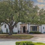 A large white two-story house with a red-tiled roof, arched windows, and a grand entryway. A large tree stands in the front yard and a curved driveway leads to the entrance. The sky is partly cloudy.