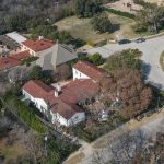 Aerial view of a neighborhood with large houses featuring red-tiled roofs, surrounded by trees and greenery, situated along a curved street with several parked cars.