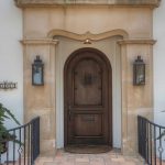 Arched wooden front door with iron accents, framed by stone columns and lanterns. Two potted topiary plants flank the entrance. A tiled walkway and wrought iron railings lead up to the door.