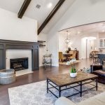 Spacious living room with exposed ceiling beams, a dark fireplace, and a cozy seating area. The room opens to a modern kitchen with pendant lights and barstools. Neutral tones and hardwood floors throughout.