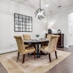 A modern dining room with a dark wooden table, four tan upholstered chairs, a beige rug, a geometric pendant light, and a mirror on the wall; hardwood floors and an arched hallway are visible in the background.