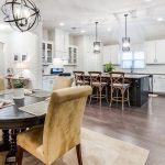 Open-concept kitchen and dining area with a round table and four tan chairs, a kitchen island with bar stools, stainless steel appliances, white cabinets, and modern pendant lighting.
