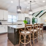 Modern open-concept kitchen and living room with white cabinets, a large island with wooden barstools, pendant lights, and hardwood floors. Living area features a sofa, plants, and large windows with ample natural light.