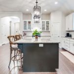 Bright, modern kitchen with a large central island, granite countertops, two wooden bar stools, white cabinets, stainless steel appliances, hardwood floors, pendant lighting, and a plant centerpiece.
