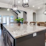 Spacious modern kitchen with a large granite island, potted plant, and pendant lights; open layout leads to a dining area with a mirrored wall and a cozy living room with a couch and large windows in the background.