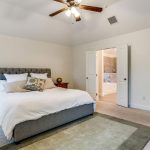 A bright bedroom with a gray upholstered bed, white bedding, pillows, a ceiling fan, and an open doorway leading to a bathroom. A green rug is on the light carpet, and a small table is beside the bed.