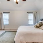 A bright bedroom with a large bed, decorative pillows, a green and beige rug, two windows with curtains, a wooden dresser, and a TV. The walls are white and the ceiling has a fan with lights.