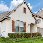 A modern, white brick house with arched windows, wooden shutters, and a two-car garage sits on a neatly trimmed green lawn under a blue sky with clouds.