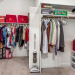 A walk-in closet with hanging clothes, storage bins, a vacuum cleaner, and shelves holding board games, pillows, and folded linens. The closet has beige carpet and white walls.