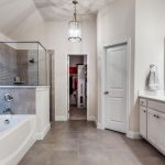 Modern bathroom with a large glass shower, white bathtub, double sinks with white cabinets, large wall mirror, and neutral-toned tile flooring. A chandelier hangs from the ceiling, and a closet is visible through an open door.