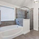 Modern bathroom with a white tub, a glass-enclosed shower, gray tile flooring and walls, a window with blinds, and an open door leading to a walk-in closet with clothes hanging inside.