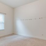A mostly empty room with beige carpet, cream-colored walls, a window with sheer curtains, and a string with small colorful decorations hanging on the wall.