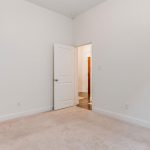 An empty, carpeted room with white walls and white doors; one door is open, leading to a hallway with wood flooring and brighter lighting.