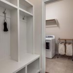 A laundry room with a washing machine, a laundry sorter with two bags, and a wall shelf, viewed through a doorway from a mudroom area with white cubbies, hooks, and a bench.
