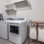 A laundry room with a white washing machine and dryer side by side, shelves above them, an ironing board leaning against the wall, and a metal laundry sorter with two fabric bags and a wooden top.