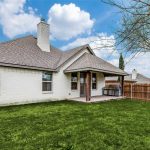 A single-story white brick house with a covered patio in the backyard, set on green grass with a wooden fence and a tree to the right. The sky is blue with scattered clouds.