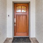 A wooden front door with a small arched window at the top, set in a white brick entryway. A decorative black doormat lies in front of the door, and a doorbell is mounted on the left wall.