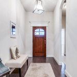 A bright hallway with a wooden front door, a beige bench with pillows, a decorative lamp on the floor, a framed picture on the wall, and a patterned rug on dark hardwood flooring.