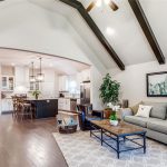 Open-concept living room and kitchen with vaulted ceiling, exposed dark beams, hardwood floors, gray sofa, coffee table, large plant, and modern decor. The kitchen features white cabinets and a central island with barstools.