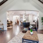 Spacious living room with vaulted ceiling, exposed beams, and hardwood floors. Features a gray sofa, coffee table, fireplace, indoor plant, and open view into a modern kitchen and dining area.